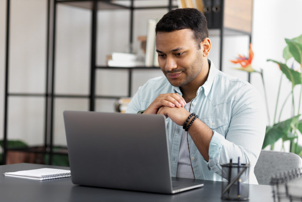 Focused millennial Indian man looking at laptop screen sitting at home office, sitting at desk with computer enjoying remote job, online studying at home concept distance education